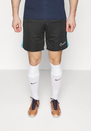 Man draagt zwarte en blauwe sportshorts, witte Nike-sokken en bronzen en zwarte sportschoenen, staand tegen een witte achtergrond.