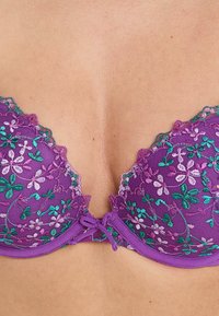 Reggiseno in pizzo viola con ricami floreali in rosa e verde. I bordi ondulati e un piccolo fiocco al centro aggiungono dettagli decorativi.