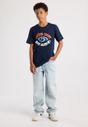 T-shirt bleu marine avec "GOLDEN COAST SURF ACADEMY" en orange et blanc, assortie avec un jean bleu clair et des baskets blanches.