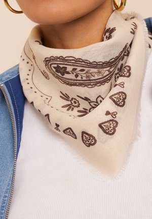 Beige sjaal met bruine bloemen- en hartpatronen om de hals geknoopt, gedragen met gouden oorringen, wit overhemd en blauwe spijkerjas.