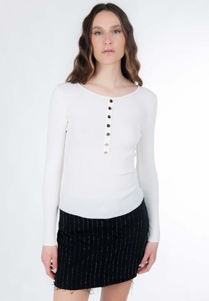 Haut blanc à manches longues avec un motif côtelé texturé, doté d'un col rond et de sept boutons sur le devant. Associé à une mini-jupe noire à franges.