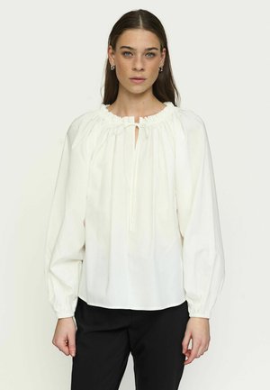 Kobieta w luźnej białej bluzce z marszczonym dekoltem i długimi, bufkowymi rękawami, w połączeniu z czarnymi spodniami, stojąca na jednolitym tle.