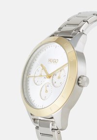 Reloj metálico plateado con acentos dorados, esfera blanca texturizada, tres subesferas y una correa de acero inoxidable con un acabado cepillado.