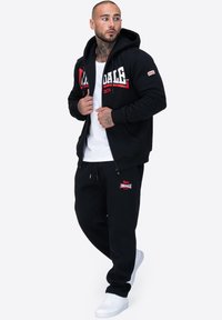 Czarny komplet bluzy i spodni dresowych wykonany z mieszanki bawełny. Bluza posiada czerwono-białe logo; spodnie mają odpowiadającą łatkę z logo. Białe sneakersy.