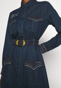 Sukienka koszulowa z dżinsu w ciemnoniebieskim kolorze z dwoma kieszeniami na piersi, haftowanymi detalami oraz szerokim paskiem z dużą okrągłą klamrą.
