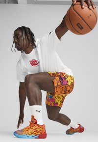 Basketbola spēlētājs baltā krekliņā, ar oranžām rakstainām šortiem un krāsainām kedām ar zilām akcentiem, driblējot bumbu.