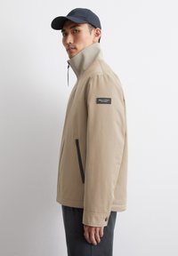 Beige Softshell-Jacke mit hohem Kragen, Reißverschluss, Seitentaschen und schwarzem Akzentlogo am linken Ärmel. Mann trägt eine dunkle Mütze.