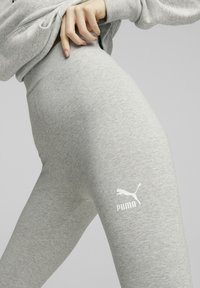 Grå leggings tillverkade av mjukt tyg, med en figurnära design och en vit Puma-logotyp på låret. Slät struktur med en åtsittande midja.