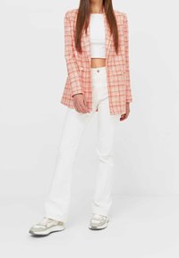 Donna che indossa un blazer a quadri arancione, un top corto bianco, pantaloni bianchi a vita alta e sneakers beige e bianche su sfondo bianco.