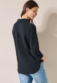 Camicetta nera in tessuto textured con colletto e vestibilità rilassata, caratterizzata da maniche a tre quarti e orlo curvo, abbinata a jeans blu chiaro.