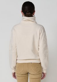 Sweatshirt crème avec un col montant, ornée de motifs floraux brodés sur les manches. Texture douce et coupe décontractée avec des poignets et un ourlet côtelés.