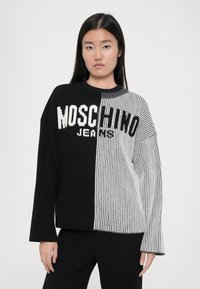 Juoda ir pilka megzta palaidinė su išilginėmis briaunomis, su drąsiu "Moschino Jeans" logotipu per visą priekį ir ilgomis rankovėmis.