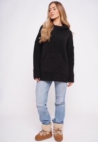 Czarny dziergany sweter z kapturem i kieszenią z przodu, zestawiony z jasnoniebieskimi dżinsami oraz brązowymi butami wyściełanymi futerkiem. Luźny fason i fakturowana tkanina.