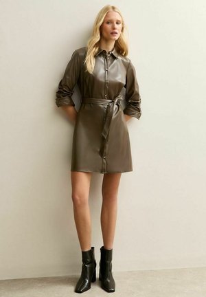 Robe chemise en simili cuir vert olive avec fermeture à boutons, poches avant et une ceinture assortie. Associée à des bottines noires.