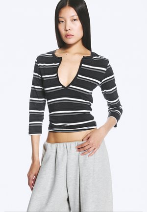 Femme aux longs cheveux noirs portant un top rayé noir et blanc à col en V profond avec des manches trois-quarts et un pantalon ample gris clair.
