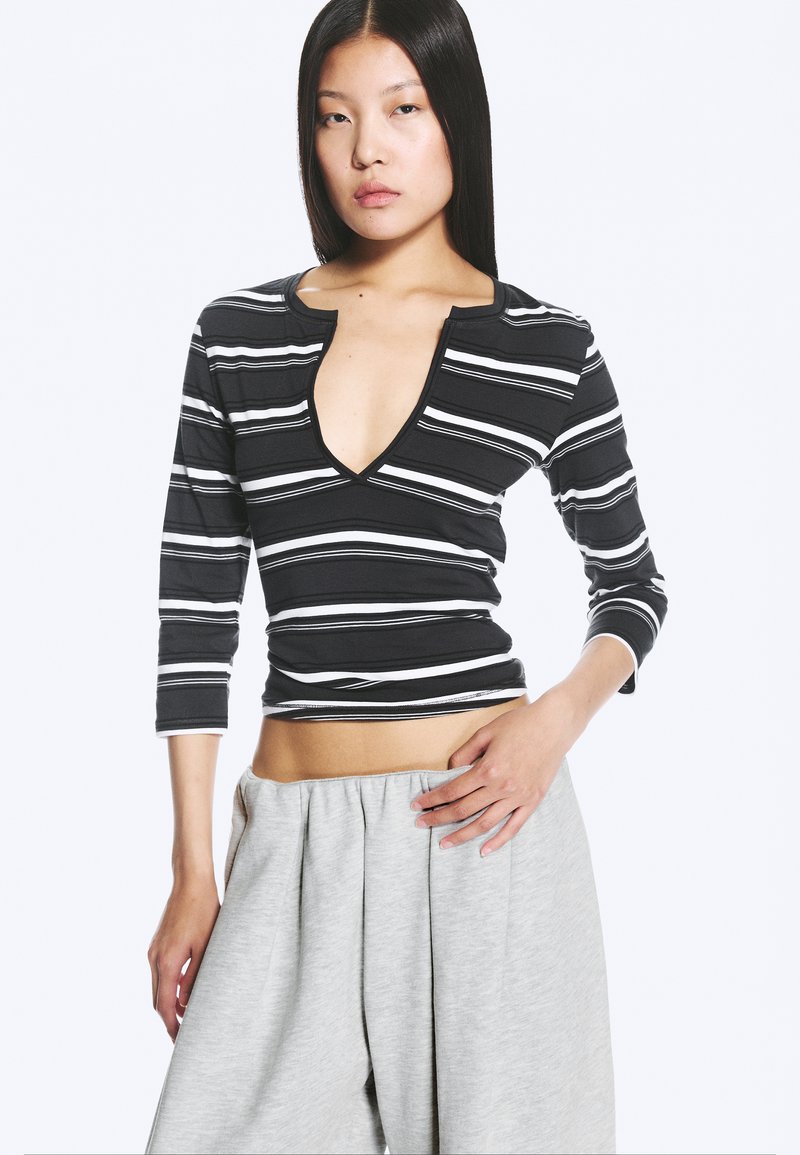 Femme aux longs cheveux noirs portant un top rayé noir et blanc à col en V profond avec des manches trois-quarts et un pantalon ample gris clair.