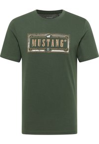 T-shirt en coton vert olive à manches courtes, arborant le logo « MUSTANG » dans un design rectangulaire avec des détails dorés vieillis et noirs.