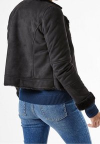 Giacca cropped nera in suede sintetico con colletto morbido in pelliccia, polsini a coste blu scuro e una texture liscia. Indossata con jeans in denim blu.