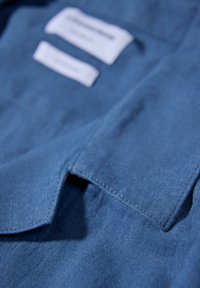 Nærbillede af stof fra blå skjorte med krave, der viser sømme og to hvide plejemærkatetiketter.