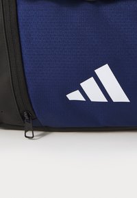 Tumesinine sportlik kott mustade aktsentidega, millel on tekstureeritud muster ja silmatorkav valge logo. Sisaldab lukuga sulgemist.