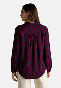 Kobieta z długimi brązowymi włosami, nosząca bluzkę z długim rękawem w czerwono-nawodnym wzorze kwiatowym oraz jasne spodnie, odwrócona tyłem.