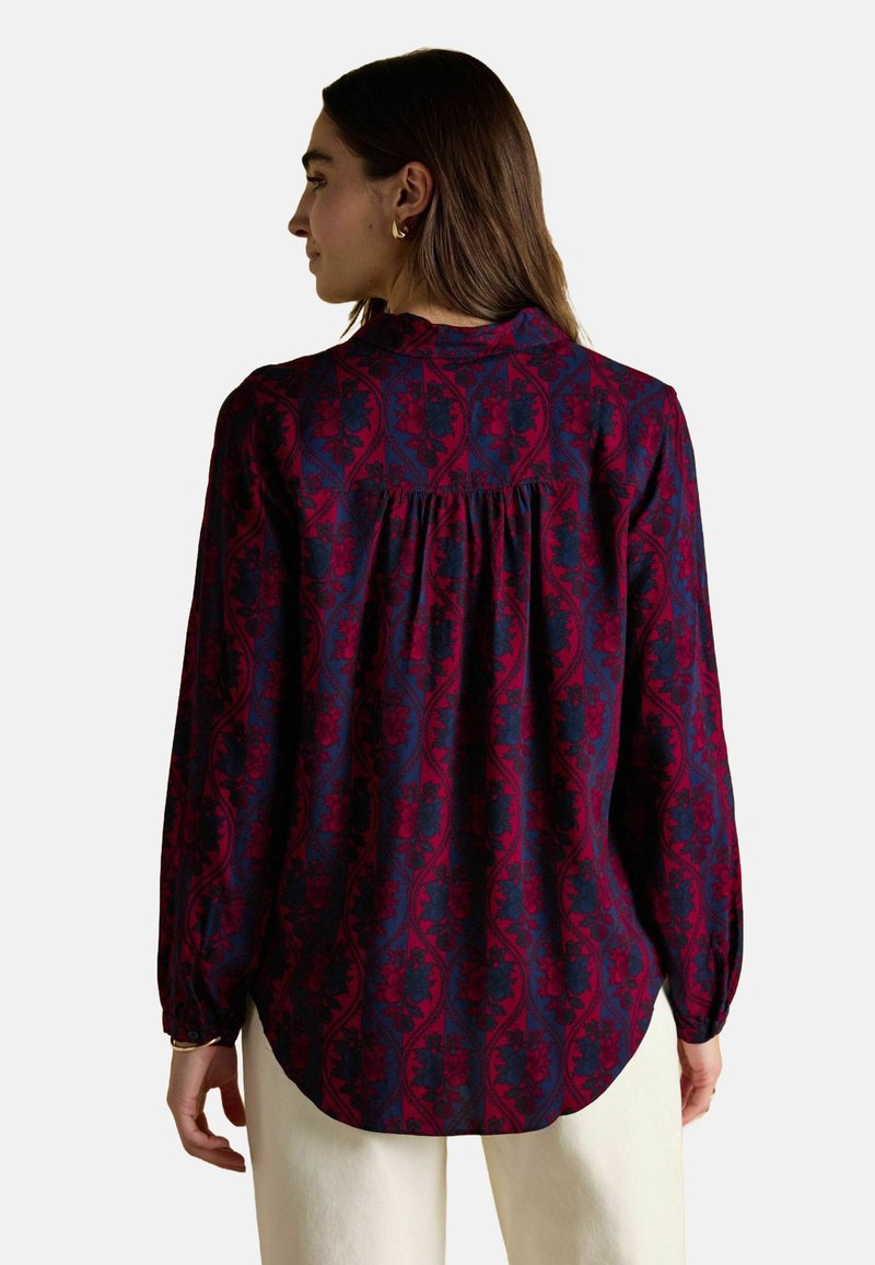 Kobieta z długimi brązowymi włosami, nosząca bluzkę z długim rękawem w czerwono-nawodnym wzorze kwiatowym oraz jasne spodnie, odwrócona tyłem.