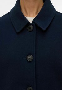 Temno modri plašč iz mešanice volne z oblikovanim ovratnikom, ki ima dva črna gumba in gladko teksturo. Pogled od blizu poudarja podrobnosti gumbov.