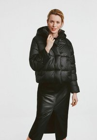 Czarna puchowa kurtka z kapturem, z pikowanym wzorem i kieszeniami z przodu, zestawiona z dopasowaną czarną skórzaną spódnicą z rozcięciem.