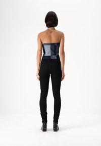 Top din denim albastru fără bretele, cu un design patchwork, asociat cu blugi skinny negri și botine cu toc negre, văzut din spate.
