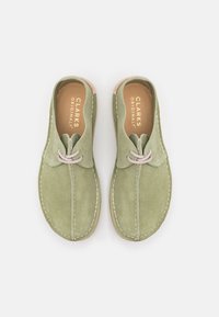 Zelené semišové boty s předním šněrováním, bílým prošíváním a hnědou podrážkou. Uvnitř je nápis "CLARKS ORIGINALS", s hladkou texturou.