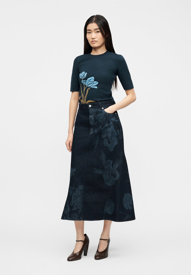 Vrouw staand met één hand in de zak, gekleed in een marineblauw topje met blauwe bloemenprint en een lange donkere denim rok met bloemmotief.