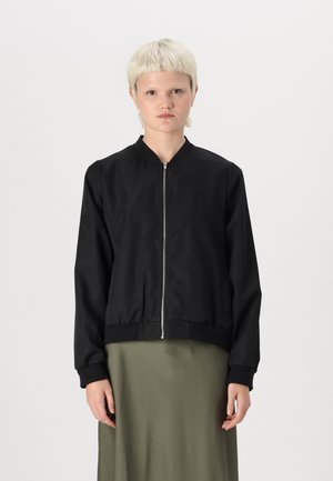 Veste bomber noire avec fermeture éclair sur le devant, poignets et ourlet côtelés. Présente une texture lisse et légèrement brillante. Portée par-dessus une jupe verte.