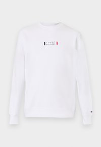 Valge dressipluus ümarate krae ja pikkade varrukatega. Rinnal on väike logo, mis on kaunistatud punaste, siniste ja valgete detailidega. Tavaline lõige.