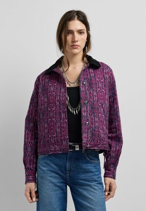 Vrouw draagt een paarse jas met patroon en zwarte kraag, gelaagde zilveren kettingen, zwart shirt en blauwe spijkerbroek tegen een effen achtergrond.