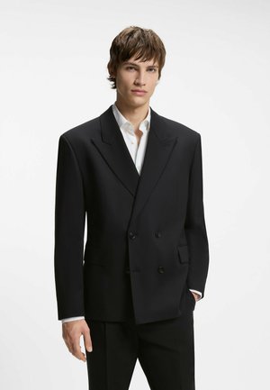 Giovane uomo che indossa una giacca da completo nera doppiopetto oversize sopra una camicia bianca, in piedi con una mano in tasca.