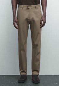 Man i slim-fit bruna byxor och mörkbruna randsydda skor, som står mot en ljusgrå vägg och ett mörkspräckligt golv.