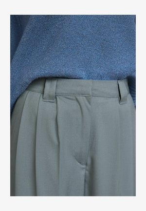 Blå stickad tröja ovanför högmidjade, veckade, ljusgrå byxor. Byxorna har en platt midja med bälteshällor och sidofickor.