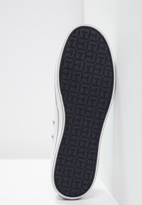 Suola di sneakers bianche rivolta in avanti con un motivo di battistrada nero che presenta forme geometriche, posizionata contro angoli di pareti bianche.