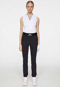 White sleeveless top with a high collar and black trim, paired with black tailored trousers and a black belt featuring a silver buckle. White glove and sneakers.