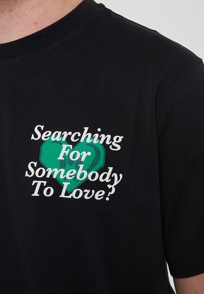 T-shirt nera con una grafica a forma di cuore verde e testo bianco che recita "Cerco qualcuno da amare?" in un font informale.