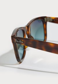 Ochelari de soare Ray-Ban cu ramă în nuanță de chihlimbar și lentile verzi închise, având un cadru robust și logo pe templu pentru branding.