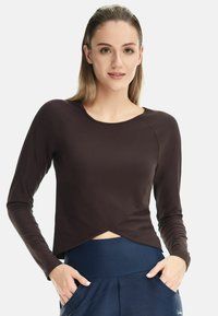 Brown long-sleeve cropped top with a rounded neckline and wrap design, paired with dark blue high-waisted pants with pockets.