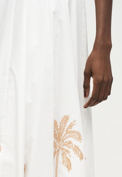 Δεξί χέρι ατόμου με σκουρόχρωμο δέρμα δίπλα σε λευκό ύφασμα με μπεζ κεντητό σχέδιο φοίνικα.