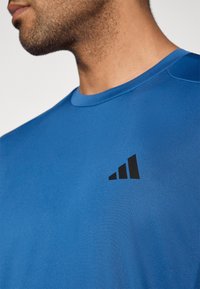 Μπλε αθλητική μπλούζα με ομαλή υφή, με μαύρο λογότυπο στην αριστερή πλευρά του στήθους. Στρογγυλή λαιμόκοψη με κοντά �μανίκια.