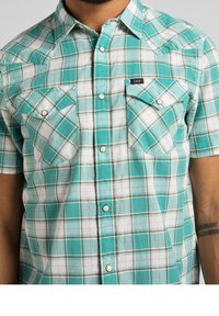 Kurzarm-Hemd im Karomuster in Petrol, Weiß und Braun. Mit zwei Brusttaschen mit Druckknopfverschluss und einem spitz zulaufenden Kragen. Aus Baumwolle.