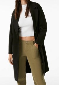 Černý dlouhý kabát s výstřihem s klopou, kombinovaný s bílým žebrovaným krátkým topem a olivově zelenými kalhotami s vysokým pasem a bočními kapsami.