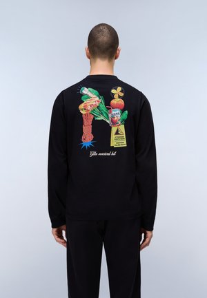 Homme portant un t-shirt à manches longues noir montrant un imprimé coloré au dos représentant une corde, un sandwich, des légumes, un carton de jus, un panneau de précaution et le texte "Kit de survie pour excursion".