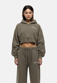 Cropped olijfgroene hoodie met een rits aan de voorkant en witte tekst, gecombineerd met bijpassende wijdvallende sweatpants met een elastische tailleband.