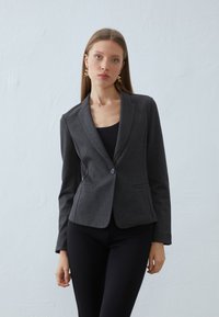 Puha anyagú, testre szabott szürke zakó, egy gombos záródással, kiemelkedő hajtókákkal és elülső zsebekkel, fekete blúzra húzva.