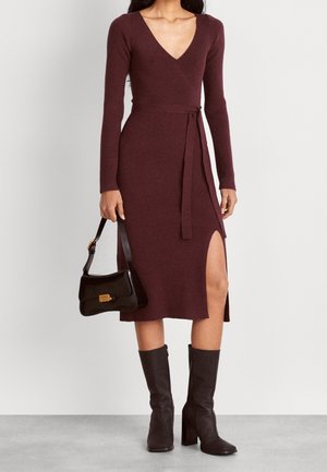 Kobieta w bordowej, dzianinowej sukience z długim rękawem, kopertowym dekoltem i rozcięciem do uda, czarnych botkach sięgających połowy łydki, trzymająca czarną torebkę.
